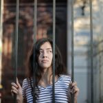 curious isolated young woman looking away through metal bars of fence with hope at entrance of modern building