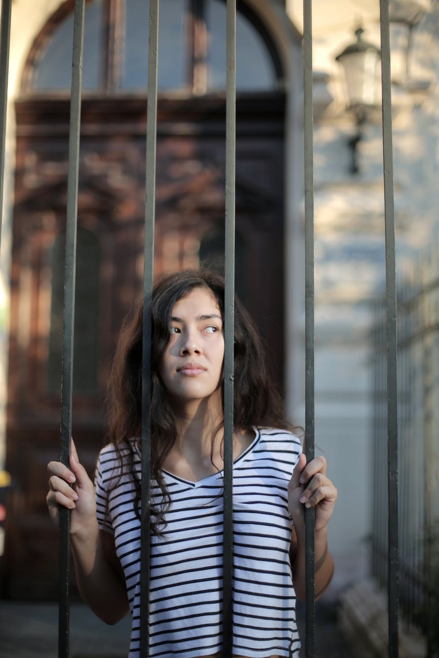 curious isolated young woman looking away through metal bars of fence with hope at entrance of modern building